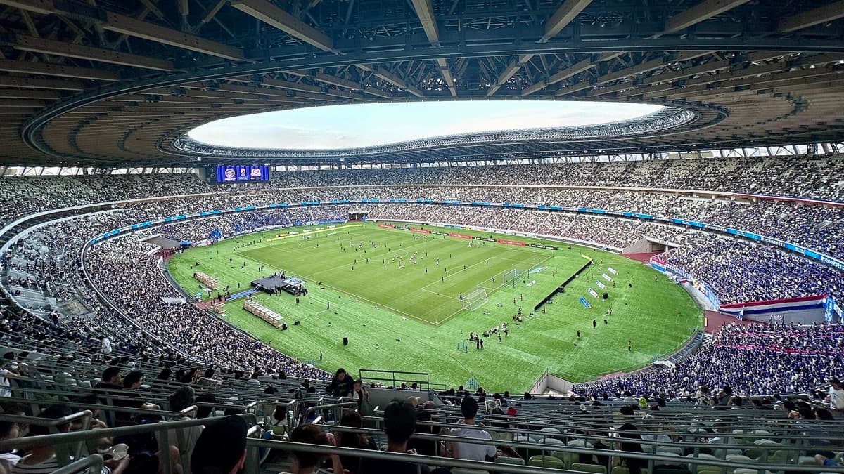 Inside a large stadium: a green pitch under an oval roof opening, with full stands all round