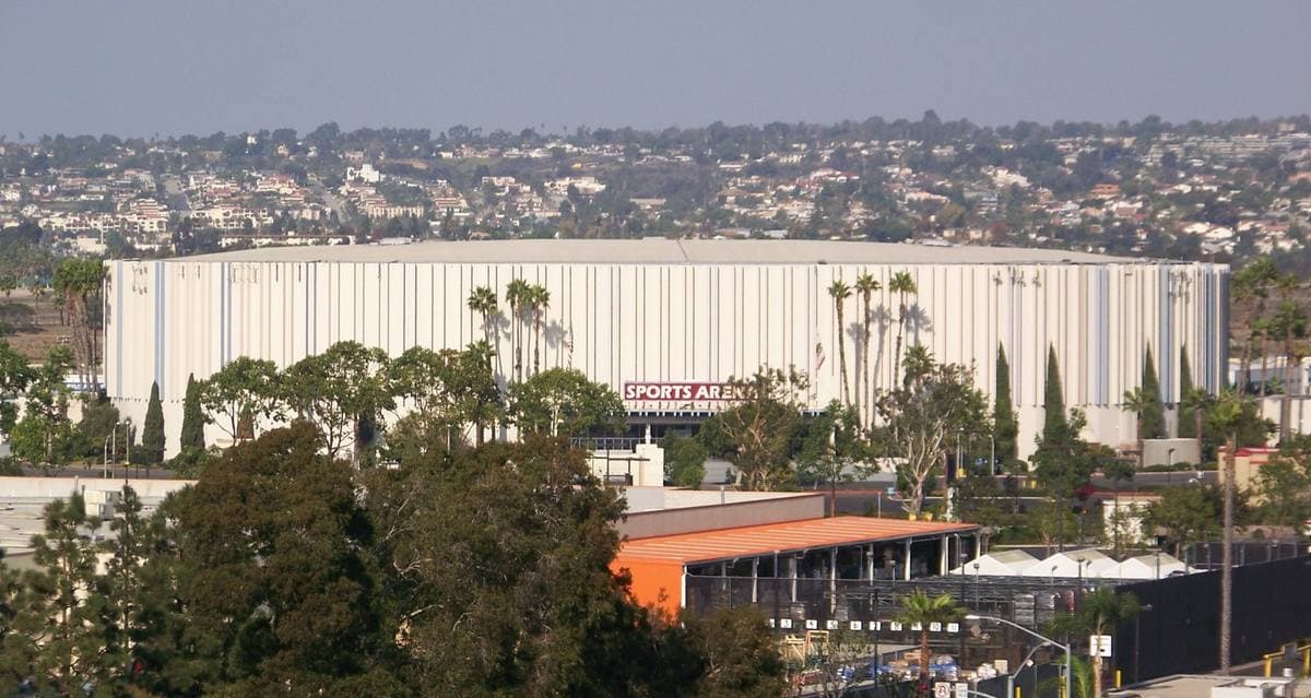 A long low white arena among trees and palms, with the hills of San Diego behind