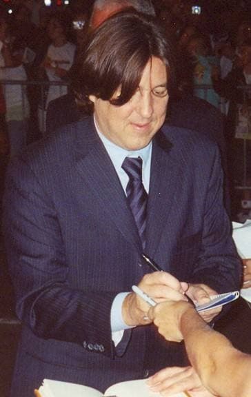 A man with a dark mop of hair and a dark suit signs an autograph