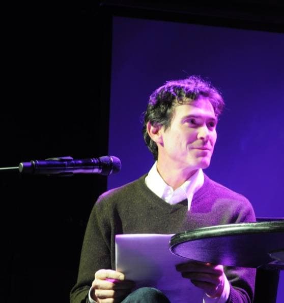A man in a dark jumper sits at a small table on a purple-lit stage, reading from a script