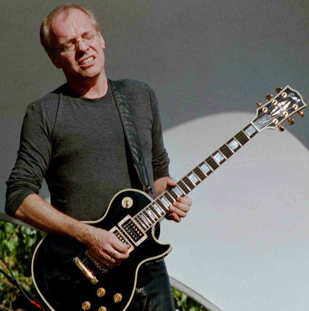 A man in a dark top plays a black Les Paul guitar on stage