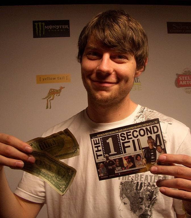 A young man with a fringe of brown hair grins and holds up a card