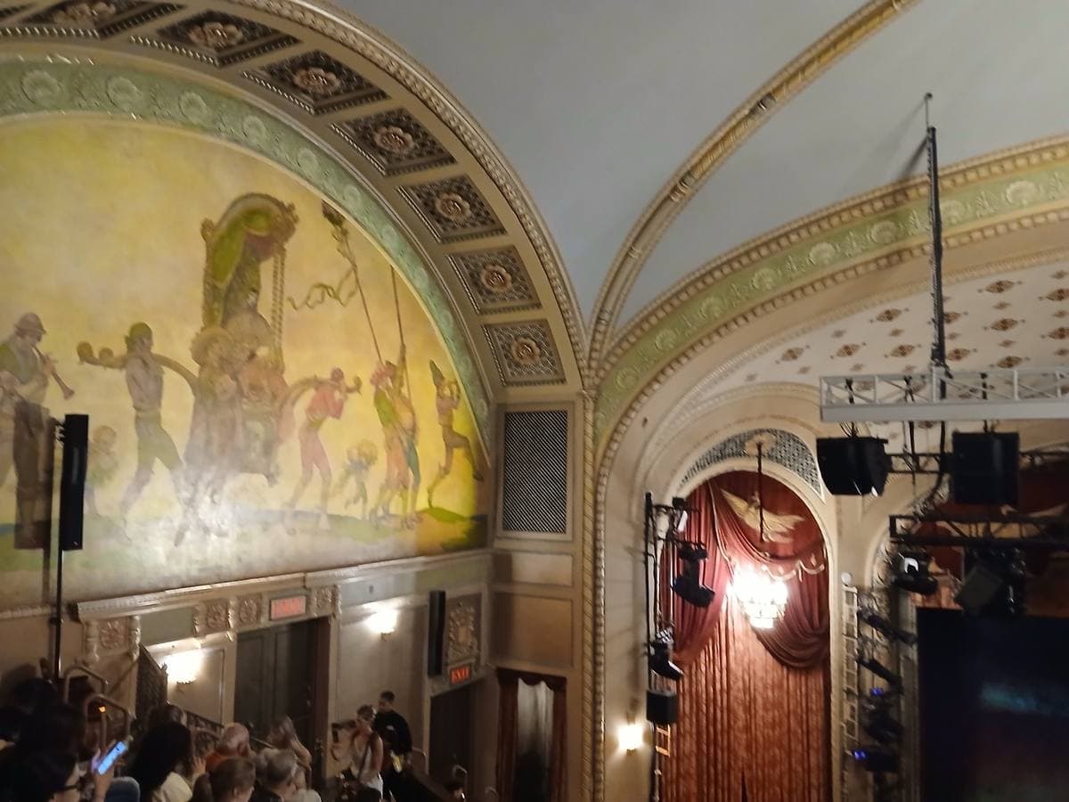 A theatre auditorium with a painted mural above the boxes and red curtains at the side of the stage