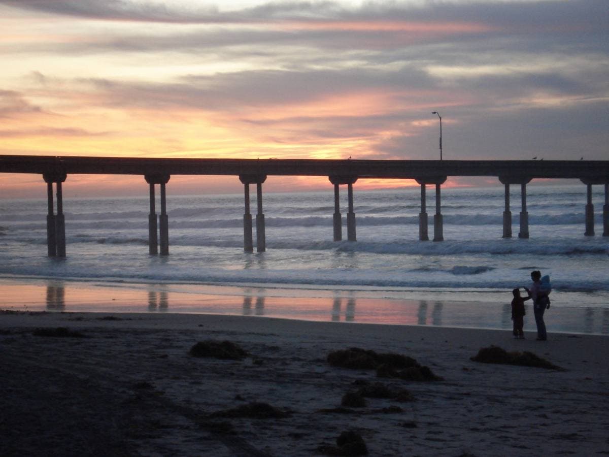 Silhouetted figures on wet sand beneath a long concrete pier at sunset
