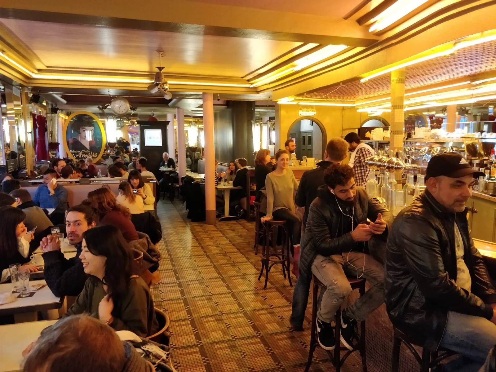 The café's busy interior: tiled floor, a long bar and customers at small tables under warm light