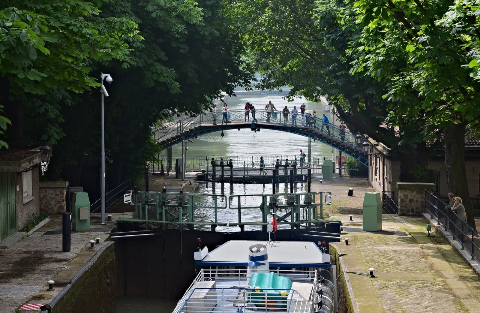 A canal lock under chestnut trees, with a tour boat waiting and an iron footbridge beyond