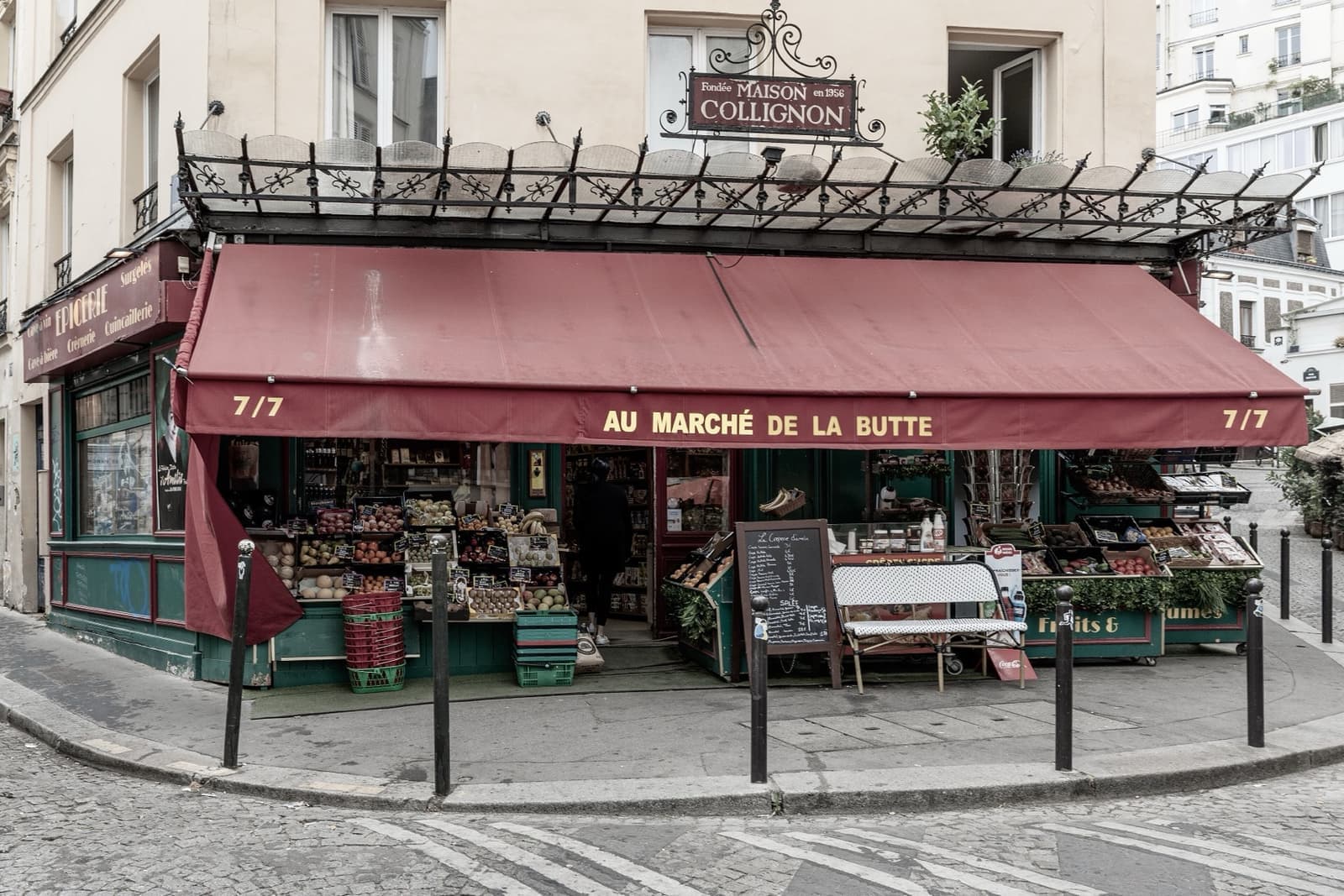 A corner grocer with a long red awning, green fruit stalls and a hanging Maison Collignon sign
