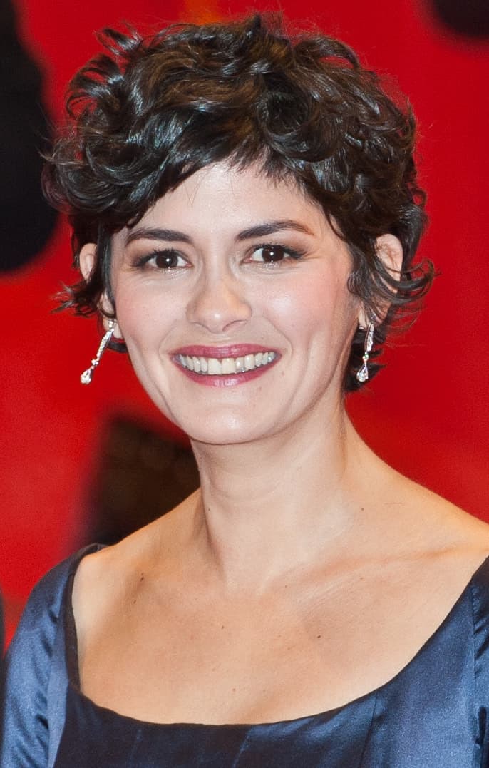 Audrey Tautou smiling, with short dark curls, against a red backdrop