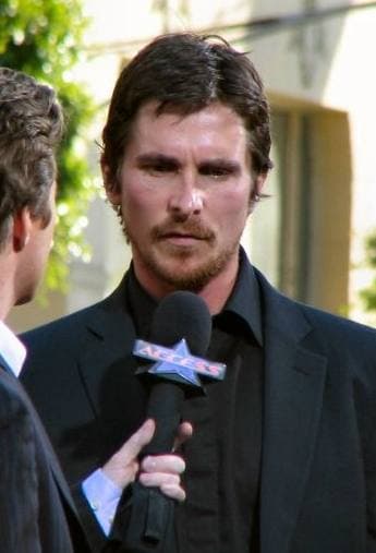Christian Bale, bearded, in a dark suit, speaking to an interviewer's microphone outdoors