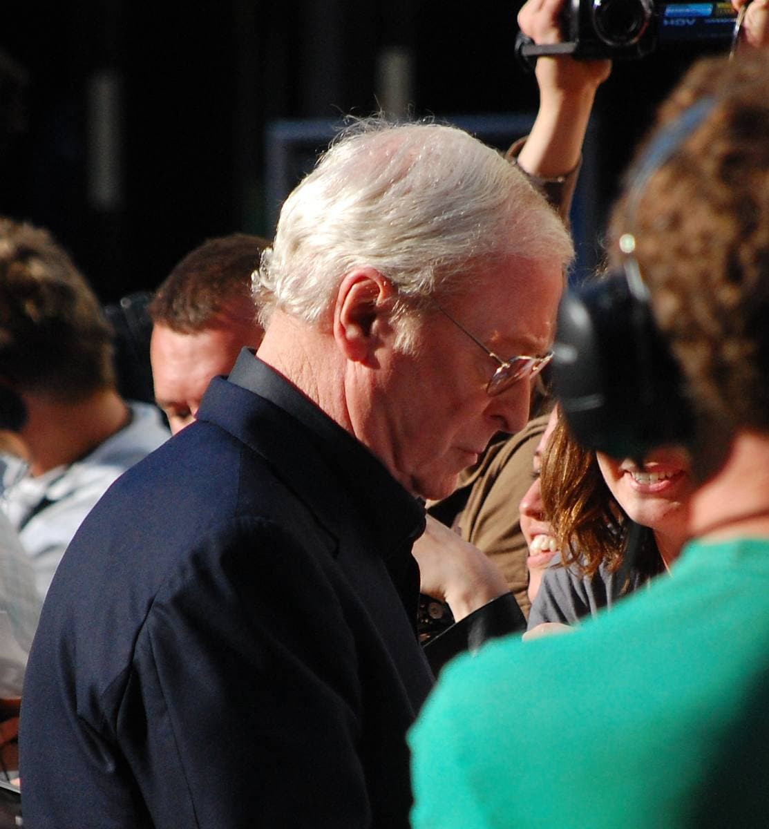 Michael Caine in glasses and a dark suit among photographers at a premiere