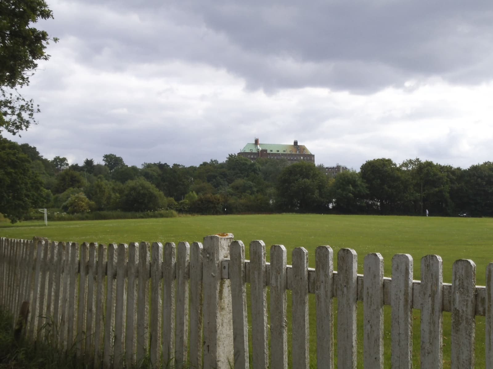 A large building with a green pitched roof rises above trees beyond a playing field