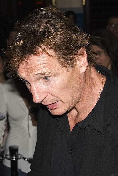 Liam Neeson in a black shirt leaning forward at a premiere