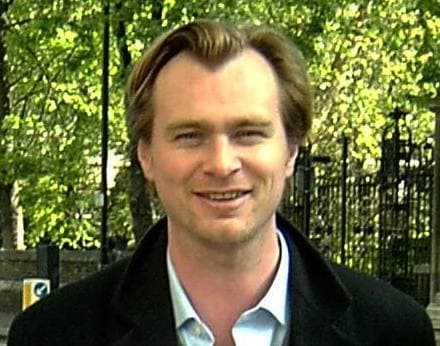 Christopher Nolan smiling in a dark coat and open-collared shirt in front of trees