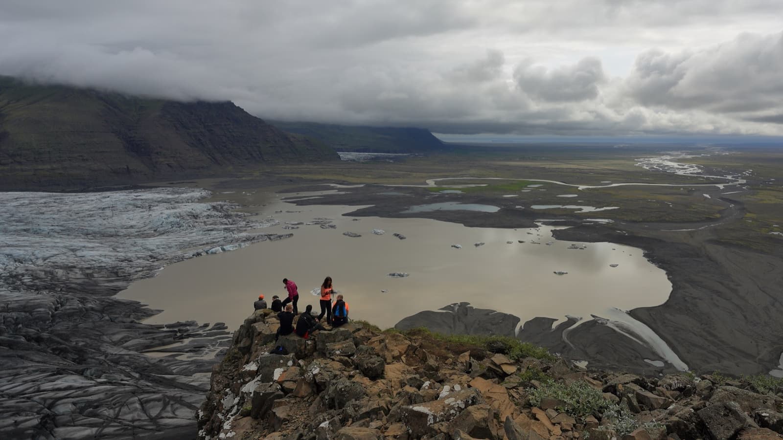 Walkers on a rocky crest look down on a glacier and its lake spreading over a dark plain