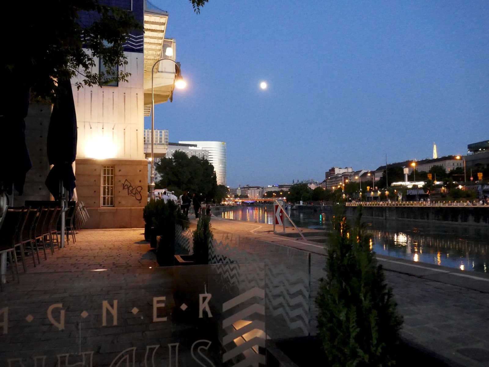 A canal bank at dusk with the moon over the water and city lights