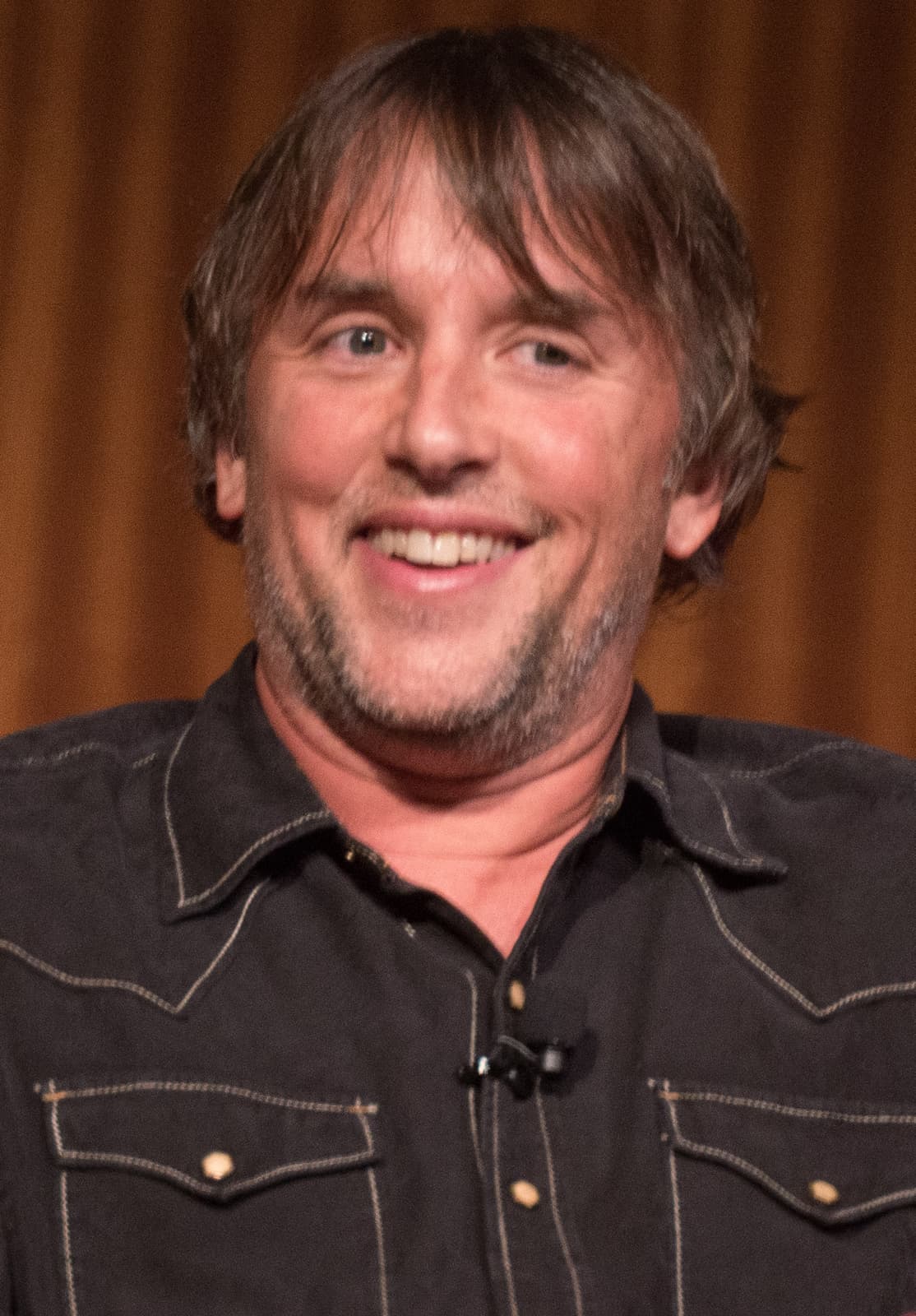 Richard Linklater smiling, in a dark western shirt, in front of a curtain