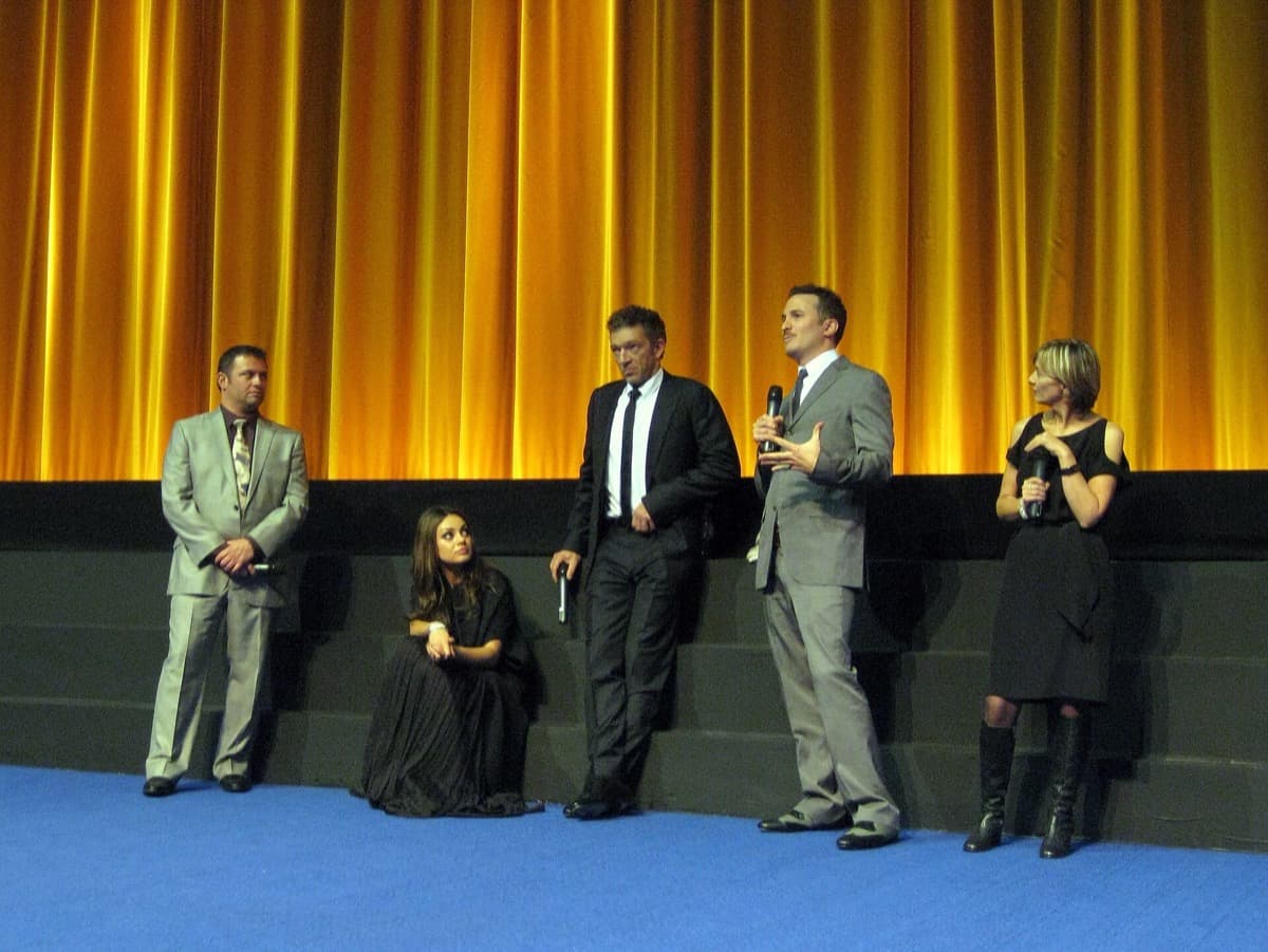 Four people and one more speaker standing and sitting before a gold stage curtain