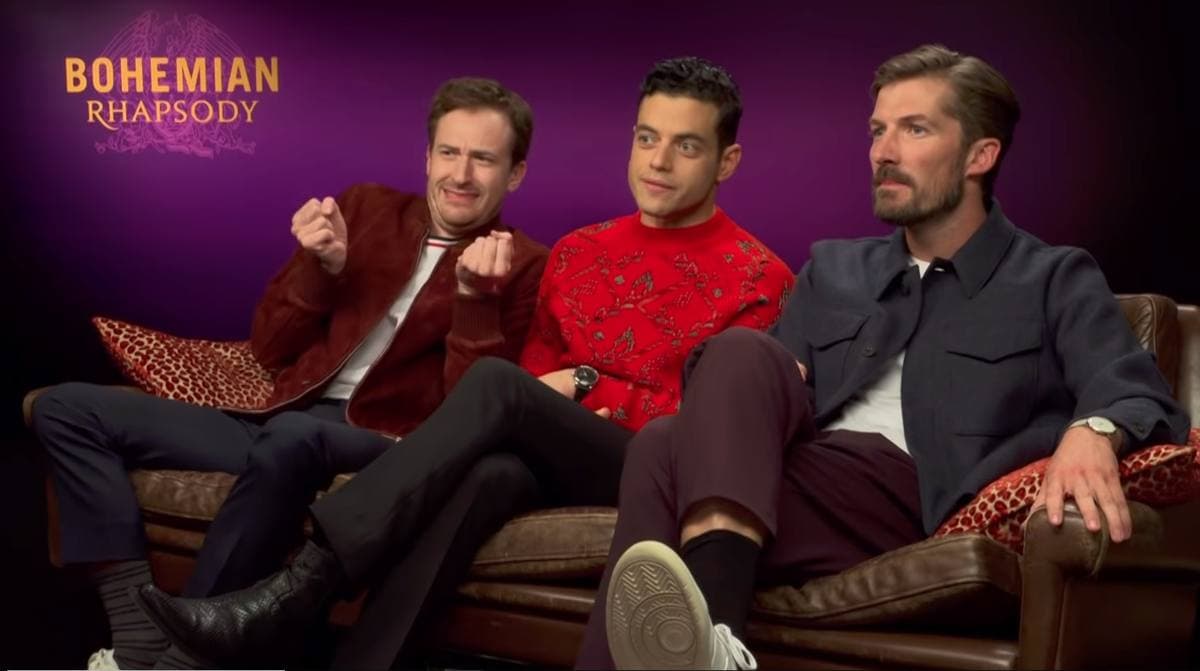Three actors on a sofa on a television set, Rami Malek in the middle in a red jumper