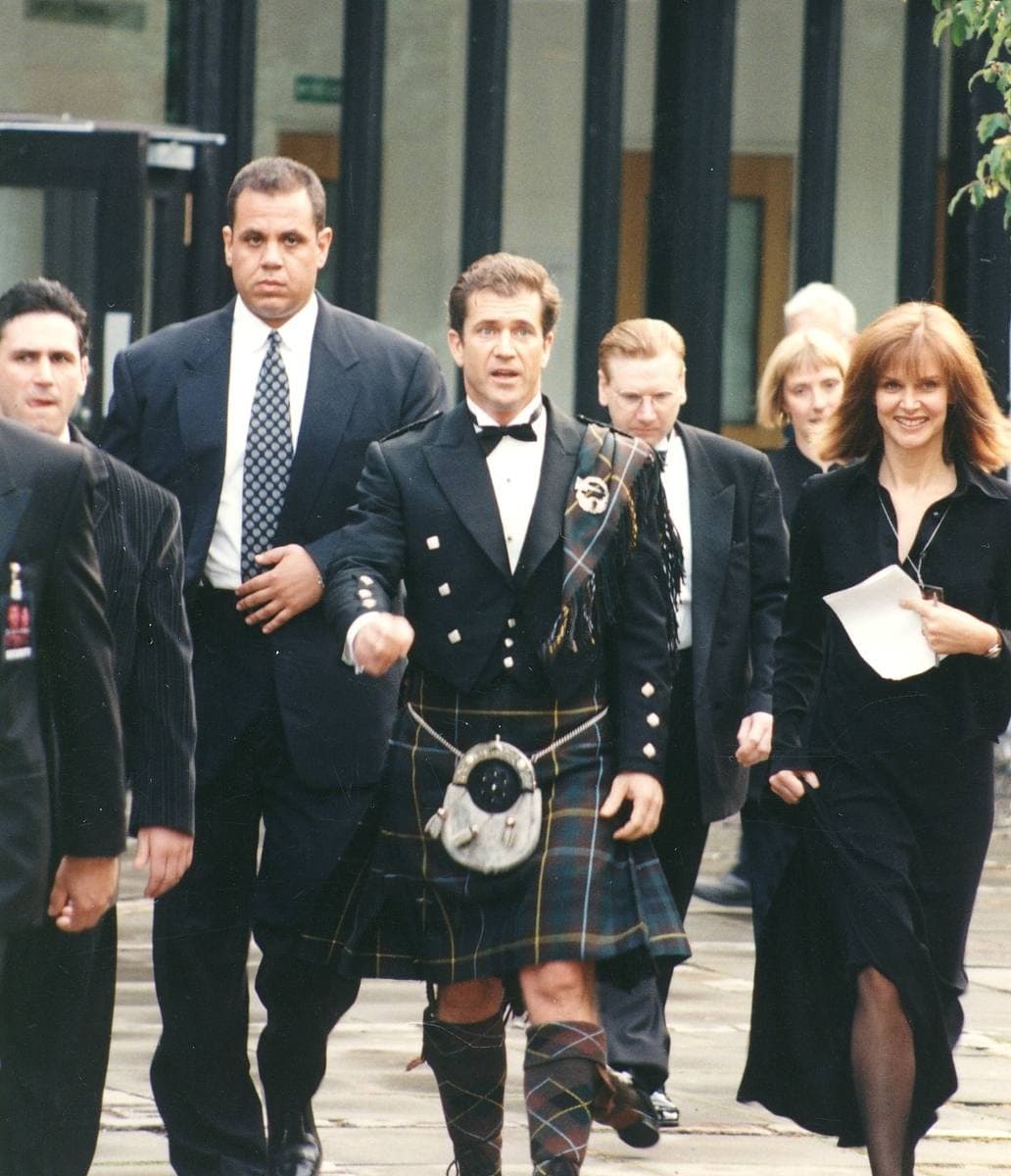 A man in Highland dress walks towards the camera among people in dark suits
