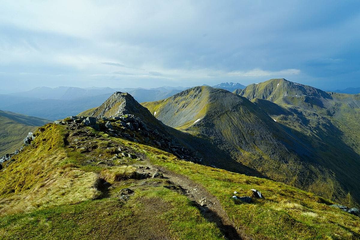 A green ridge path leading to pointed mountain summits under a pale sky