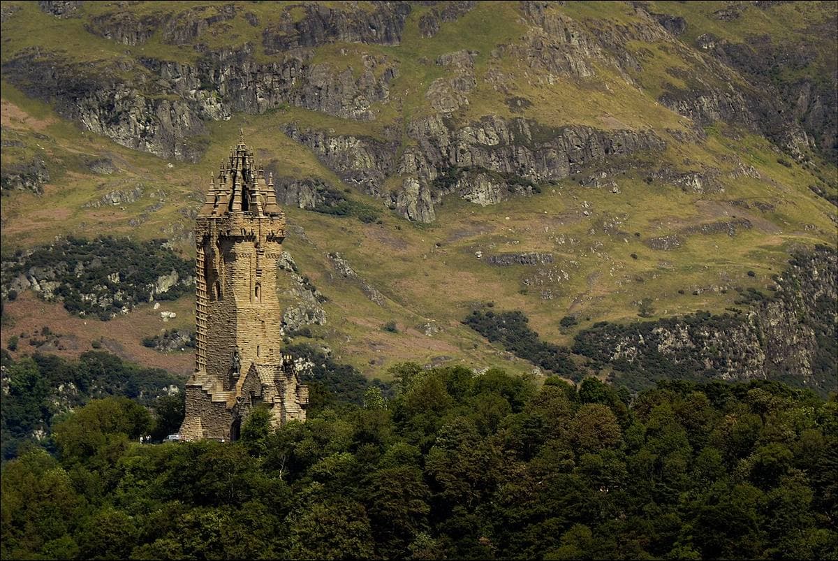 A tall stone tower with a crown-shaped top rises from trees in front of a rocky hillside