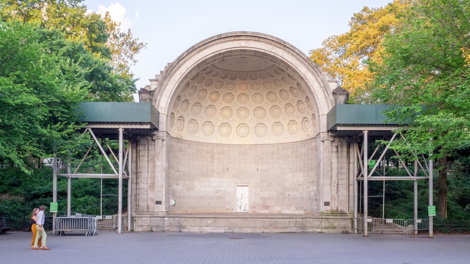 A neoclassical stone half-dome bandshell facing an open paved concert ground, trees either side