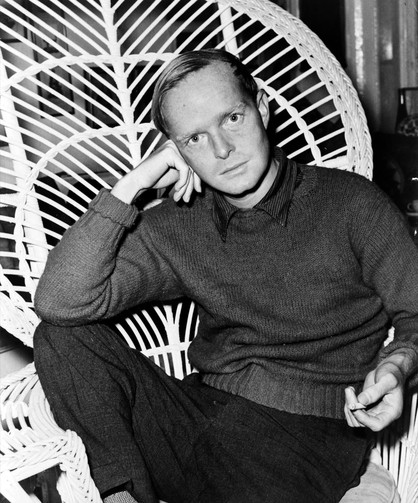 Truman Capote in a dark sweater, seated in a white wicker chair, head resting on his hand