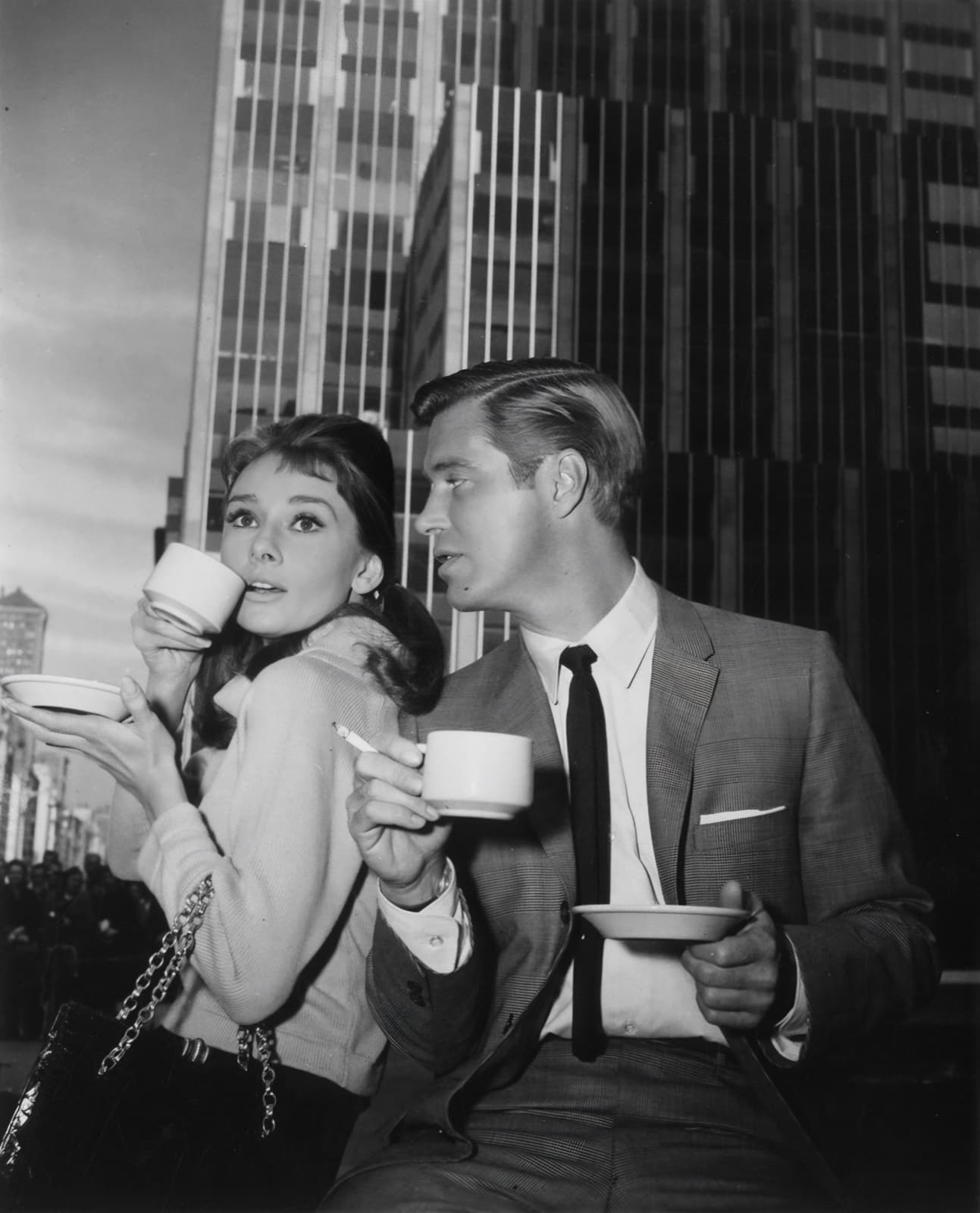 Audrey Hepburn and George Peppard standing side by side drinking from coffee cups, office towers behind