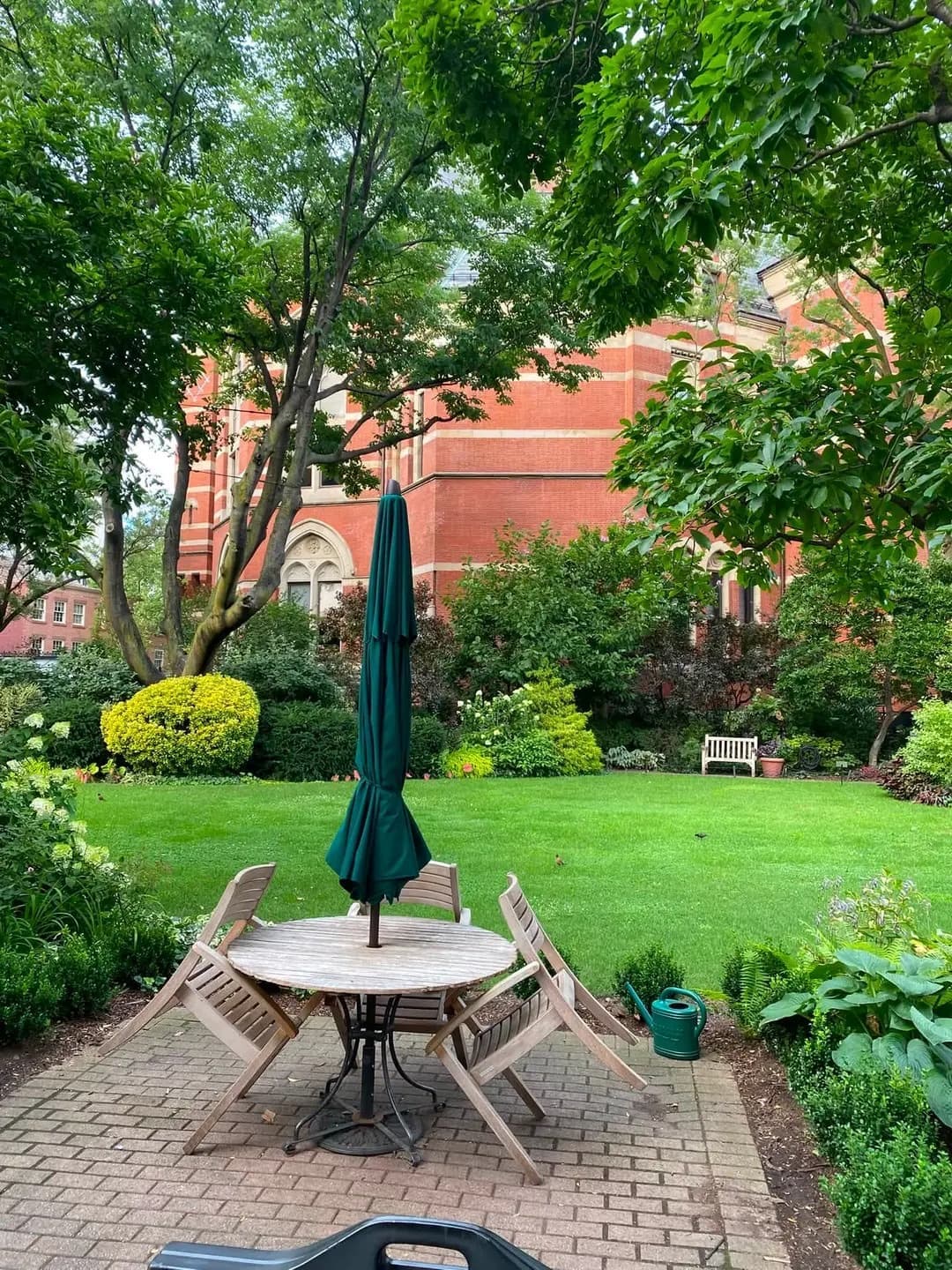 A small lawn with a table and furled green umbrella, flower beds and a red-brick Victorian building behind