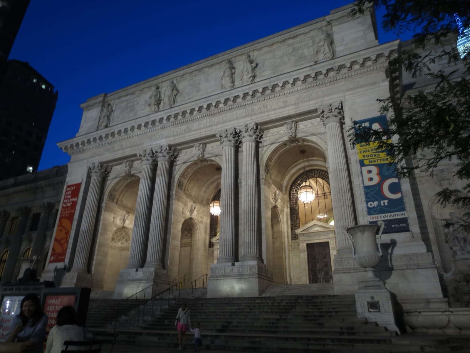 The marble Beaux-Arts front of the library, lit at dusk under a deep blue sky, with banners between the columns