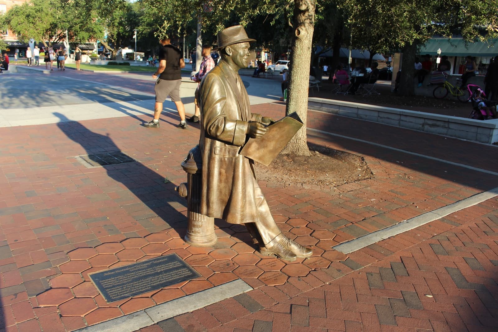 A life-size bronze of a man in a hat and overcoat leaning on a post with a newspaper, on a brick plaza