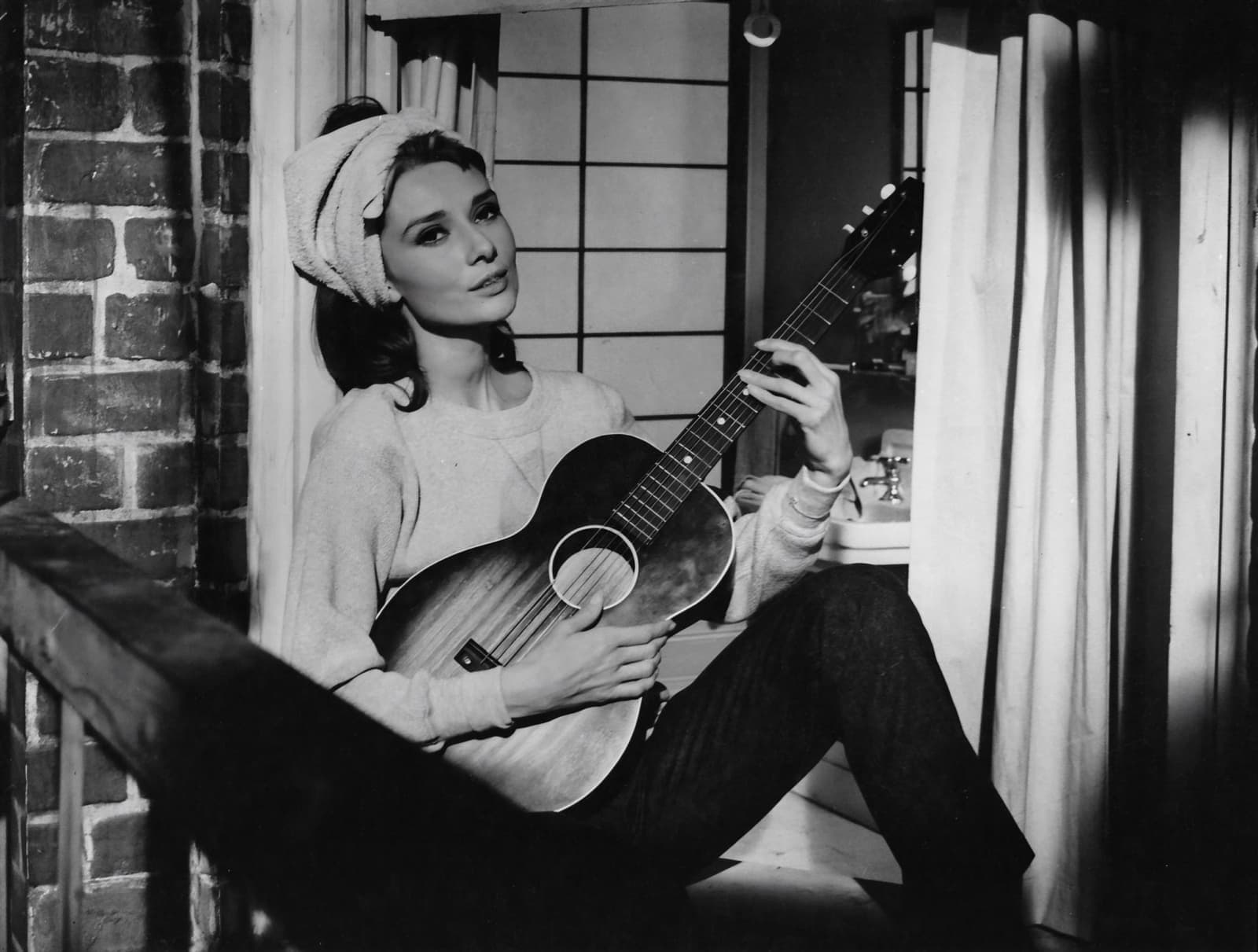 Audrey Hepburn in a sweater and headscarf sitting on a window ledge with an acoustic guitar
