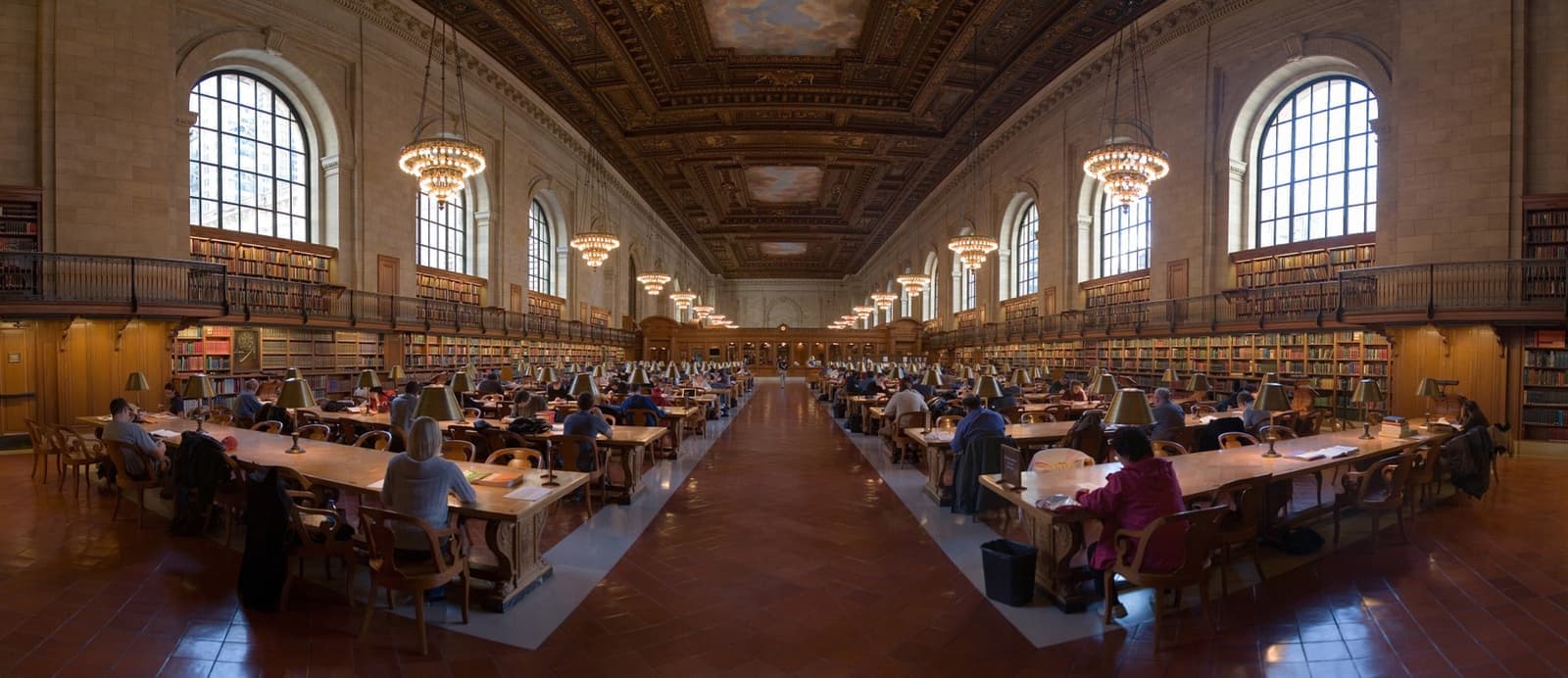 A long reading room with arched windows, chandeliers and rows of wooden tables with readers