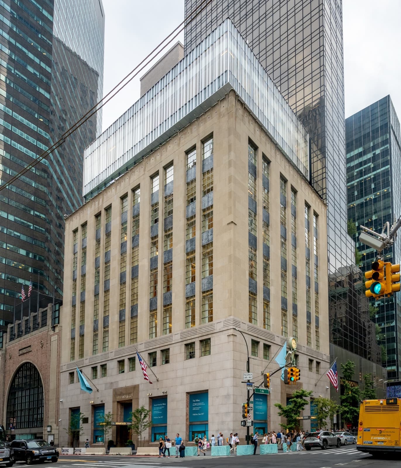 A pale limestone corner building with a glass addition on top, flags over the entrance and pedestrians on Fifth Avenue
