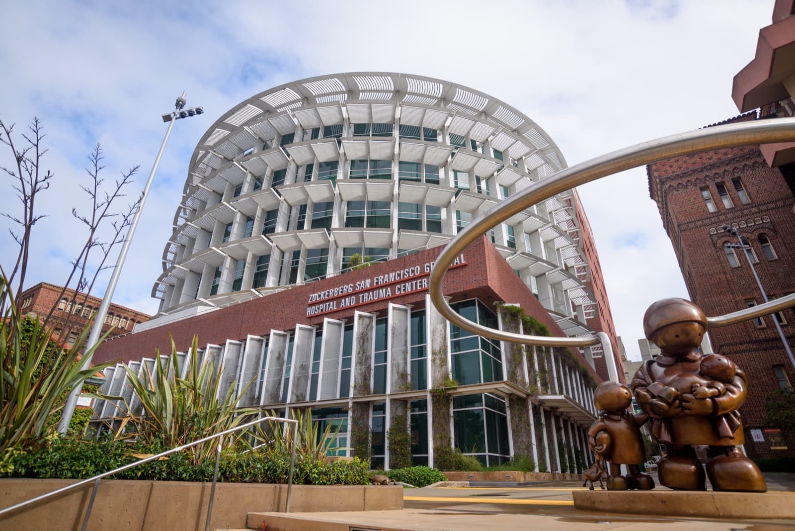 A modern hospital building of brick-red panels and glass, with a sculpture in front