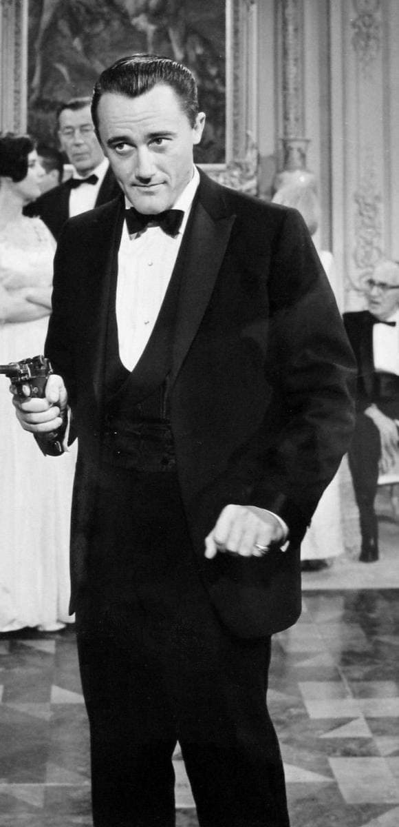 Black-and-white photograph of Robert Vaughn in a dinner jacket, holding a pistol, in a ballroom