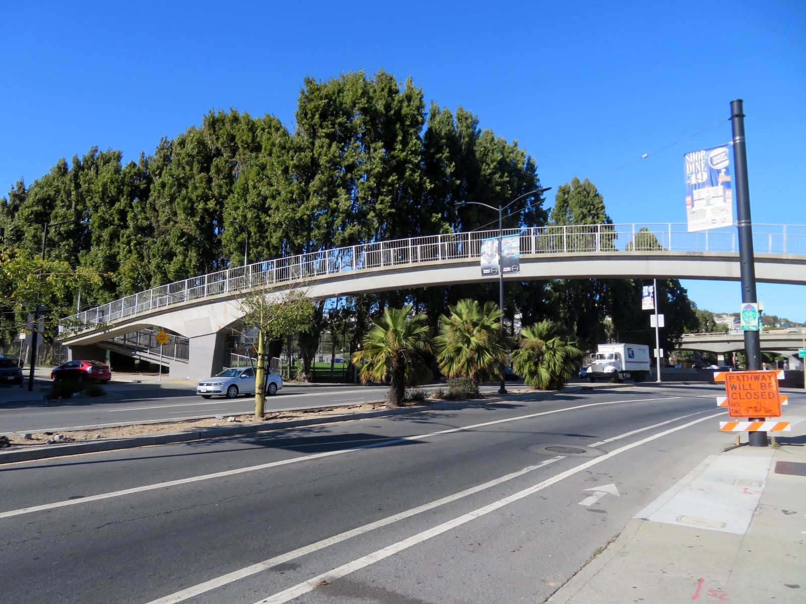 A wide road under a footbridge, with houses rising on a hill behind