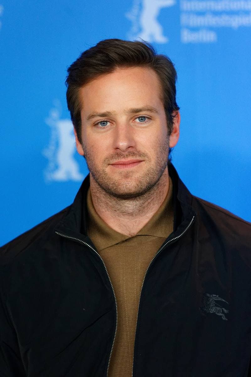 Armie Hammer in a dark jacket over a brown knit shirt, against a blue backdrop