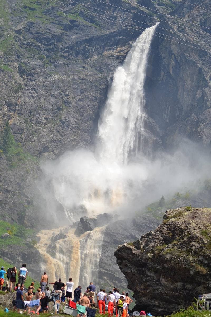 A tall white waterfall pouring down a rock face, with a crowd watching from the meadow below
