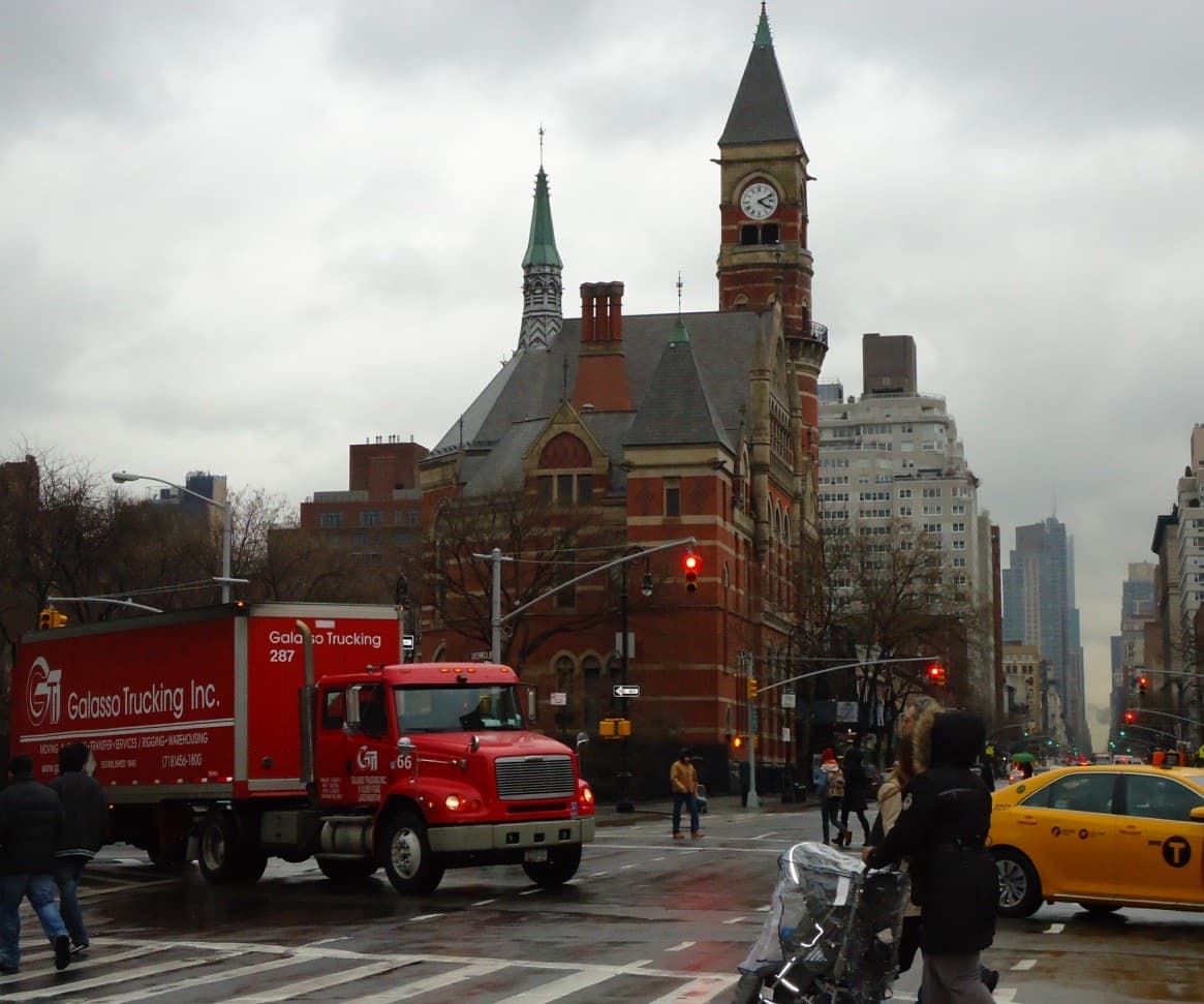 A Manhattan avenue with a Gothic brick tower rising behind street traffic
