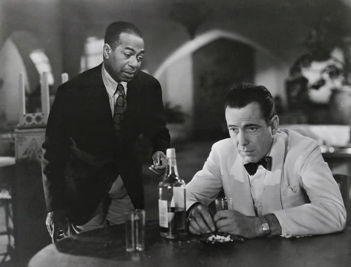 Dooley Wilson leans over a table where Humphrey Bogart sits with a bottle and glass, Moorish arches behind