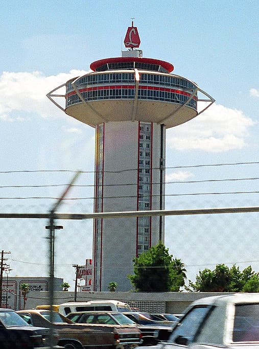 A tall white tower topped by a wide, round, saucer-shaped upper level, seen over parked cars