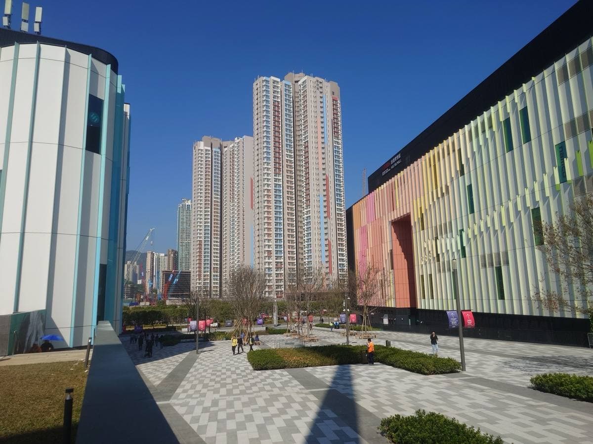 A paved plaza between the new Kai Tak Sports Park buildings and tower blocks under a blue sky