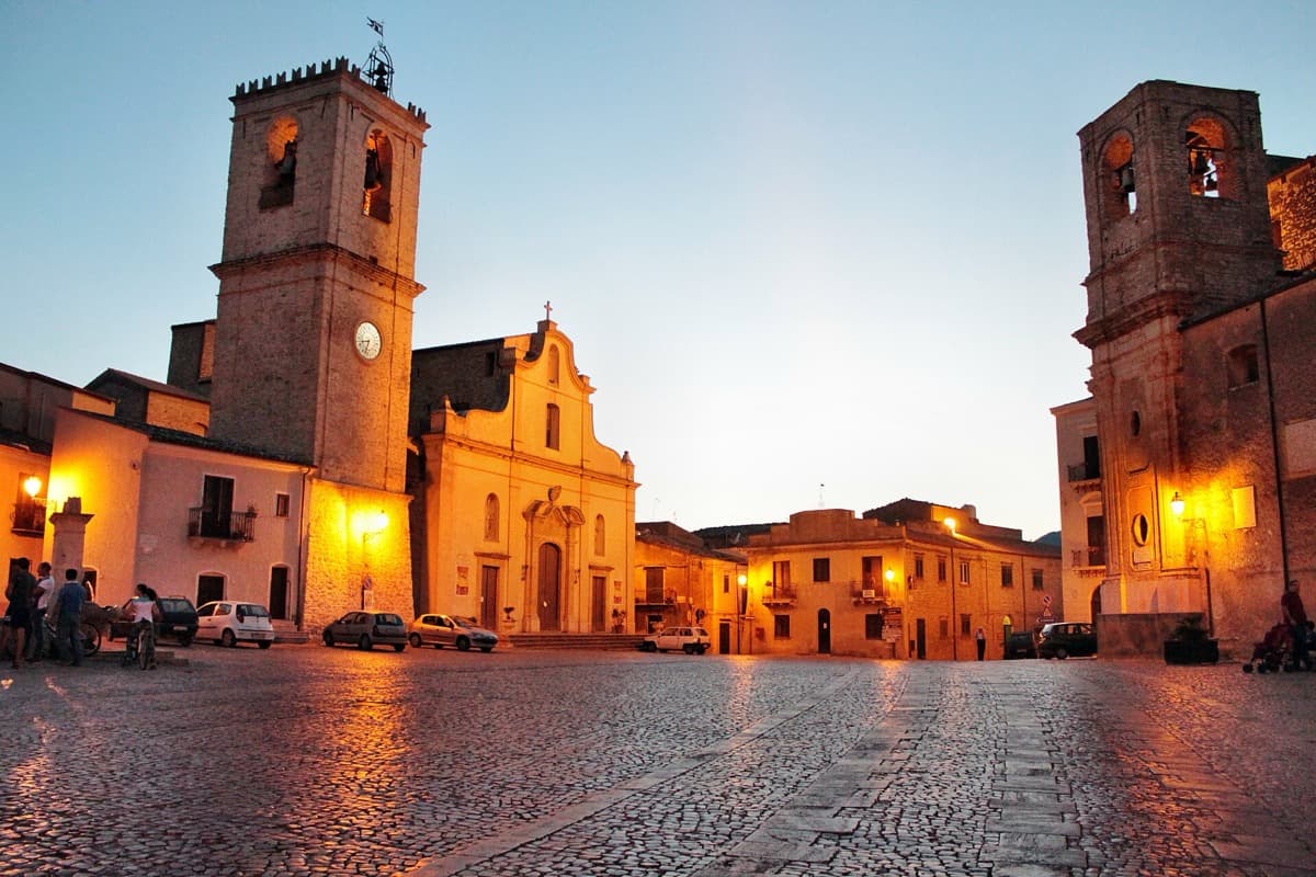 A sunlit Sicilian town square with a fountain, a church and balconied houses