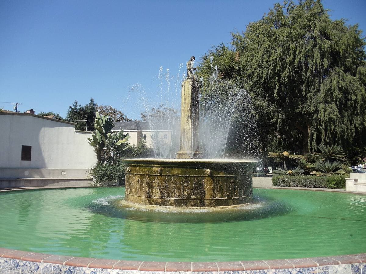 A round fountain with a green pool and a central column spraying water, trees behind