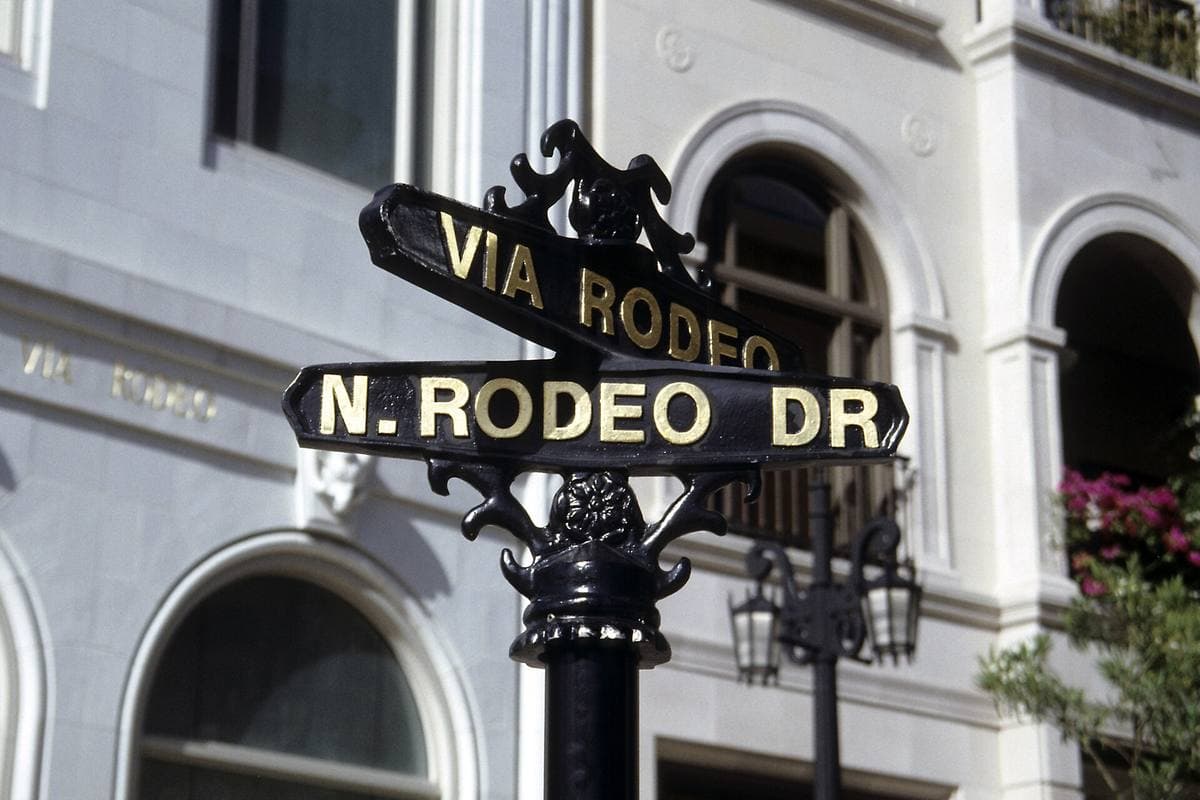 A black ornate street sign reading Via Rodeo and N. Rodeo Dr in front of a white building