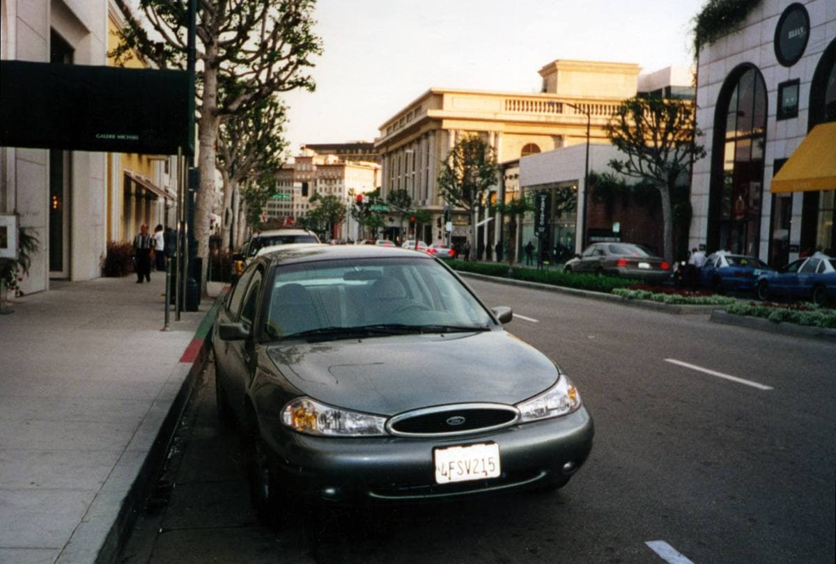 A street of low shopfronts and palm trees with a parked silver car