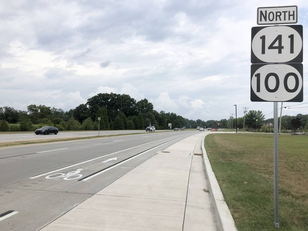 A suburban highway with route signs for 141 and 100 under a summer sky