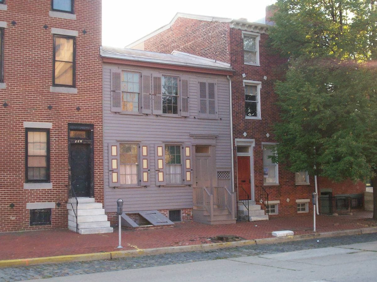 A narrow grey clapboard house between two brick houses on a city street