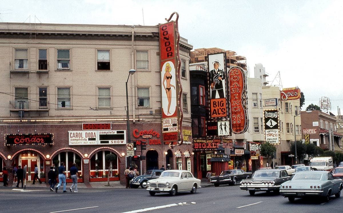 A row of strip-club fronts with tall blade signs, 1970s cars on the street